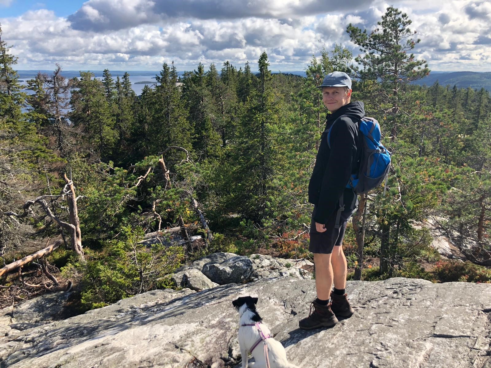 Aikuinen partiolainen Eerik Klemetti kalliolla metsämaiseman edessä koiran kanssa
