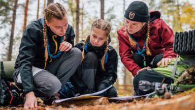 Kolme lettipäistä partiolaista katsoo yhteistä paperia luonnossa