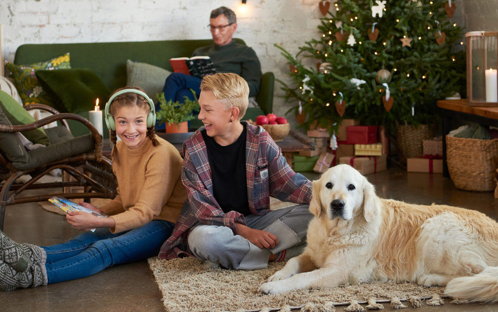Kaksi lasta istuu lattialla koiran kanssa. Toisella on päässään kuulokkeet ja kädessään partiolaisten adventtikalenteri. Taustalla vanhempi mies sohvalla ja joulukuusi.