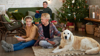 Kaksi lasta istuu lattialla koiran kanssa. Toisella on päässään kuulokkeet ja kädessään partiolaisten adventtikalenteri. Taustalla vanhempi mies sohvalla ja joulukuusi.