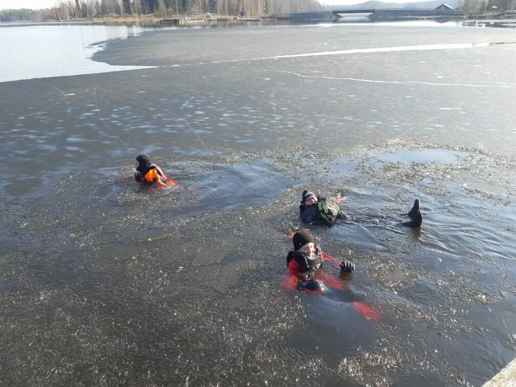 Kolme henkilöä märkäpuvuissa ja pelastusliiveissä jäisessä vedessä.